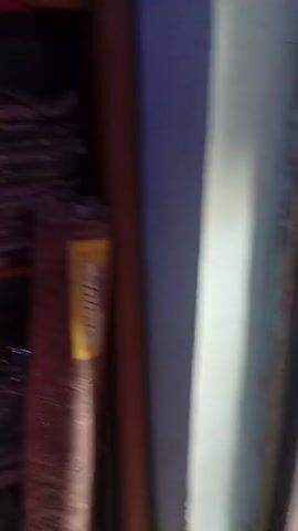 The video shows a storage shelf filled with various fabrics, including stacks of colorful clothes, in what appears to be a shop. A man in a dark tunic is seen standing nearby. The camera then pans to a collection of curtains in different colors and patterns, hanging on a rail. A wooden bench is visible in the foreground.
