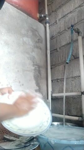 The video shows a person washing dishes in a cluttered outdoor area. The subject, whose face is not clearly visible, is wearing a black top and has their hands submerged in a blue basin filled with soapy water. They are holding a white plate, rinsing it under a stream of water coming from a blue hose. The hose is connected to a pipe system with a faucet. In the background, there is a large concrete wall, a red water tank, and a partially visible plastic basket with kitchen utensils. The ground is covered with rough bricks. The scene appears to be taking place during the day, though no specific time or weather conditions can be determined. The overall atmosphere is utilitarian, suggesting a simple, everyday chore. There is no visible text in the video. The location is identified as Arjasa, Indonesia.