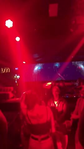 The video takes place inside a nightclub with red lighting. Initially, the focus is on the dance floor with people dancing and a large screen in the background displaying colorful graphics. The camera then moves towards a woman holding a drink who smiles at the camera. The camera blurs briefly before moving on to show the woman and a man, who is wearing a black shirt with white stripes.