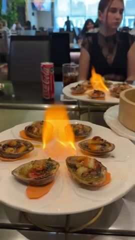The video shows a table at a restaurant. In the foreground, on a white plate, are several cooked dishes with flames igniting them. These dishes appear to be seafood, possibly escargots, served in their shells on slices of carrot. There is also a can of Coca-Cola and a glass of dark liquid on the table. In the background, a young woman in a black lace dress is seated at the table. Other patrons and restaurant staff are visible in the blurred background. The environment appears to be a dimly lit, upscale dining establishment with greenery on the walls. The camera movement is primarily focused on the flaming food.