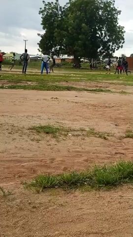 The video shows a group of young people playing soccer on a dirt field. The field is surrounded by grass and trees, with a few buildings in the background. The sky is overcast. The players are wearing casual clothing. Some are actively playing, while others are observing from the sidelines. The general atmosphere appears to be one of informal recreation.