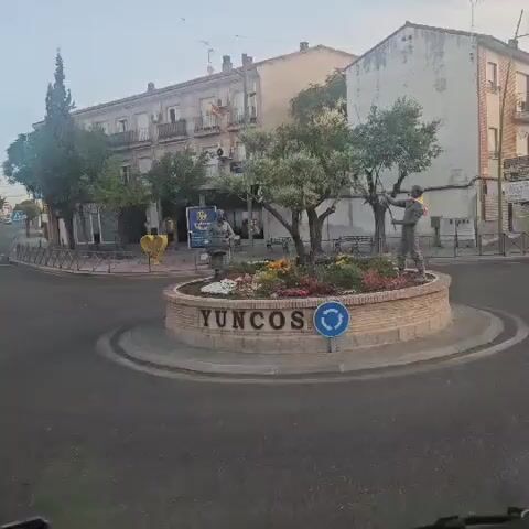 The video shows a roundabout with a circular planter in the center. The planter has flowers and two statues. One statue is of a person holding a basket, and the other is of a person holding a flag. The word YUNCOS is written on the side of the planter. There is a blue traffic sign with a white circular arrow on the side of the planter. Buildings are visible in the background.