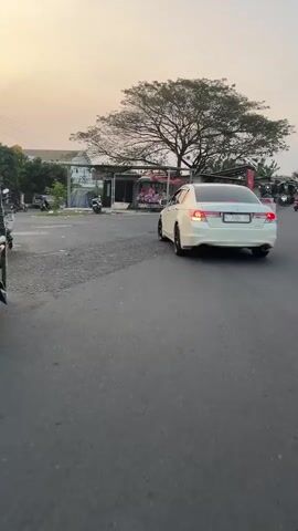 A white Honda Accord sedan is shown from behind as it drives away on an asphalt road. The car has tinted windows and a license plate that is partially visible. The road appears to be in an outdoor setting with some buildings and trees in the background. The sky is a light orange color, suggesting it is either sunrise or sunset. There are also other vehicles and people visible in the background, but they are out of focus. The car's brake lights are illuminated, and it appears to be turning slightly to the left. The overall scene is a still image, not a video.
