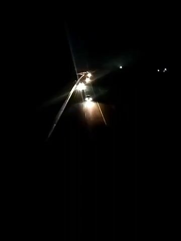 The video shows a dark night road with several cars driving. The camera is positioned behind a car, capturing its headlights and the headlights of the cars ahead. The road appears to be a highway with white lines marking the lanes. The environment is completely dark except for the illumination from the car headlights. The cars are moving in the same direction.