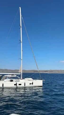 A white sailboat is shown from its starboard side, floating stationary on blue, rippling water under a clear, bright blue sky. The boat has a white hull, a tall white mast, and a boom. Distant land with hills is visible on the horizon. No people are visible on the boat.