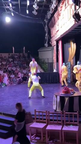 The video shows a performance on a stage in front of an audience. In the center of the stage, two performers in yellow outfits are engaged in a balancing act. One performer is standing on the shoulders of the other, and the top performer briefly jumps into the air. To the right of the stage, three performers in yellow suits with blue masks are standing. A round table with a cake and fruit is placed to the right of the performers. The stage is lit with purple lights, and the audience is seated in tiered seating in the background. Two empty chairs are in the foreground.