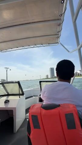 A person is seen from behind, steering a small motorboat on a sunny day. The individual is wearing a white or light-colored shirt and a bright orange life vest, seated at the helm with their back to the camera. The boat itself is white, with a black seat and a white canopy supported by metal frames, providing shade from the bright sun.

The boat is on calm, blue water, with a clear sky overhead. In the distance, a modern cityscape is visible, characterized by several tall, high-rise buildings. Stretching across the background, a prominent cable car system can be seen, with multiple towers extending over the water or along the shoreline, suggesting a developed urban or tourist destination. The scene appears to depict a leisure activity, possibly a scenic boat ride or transit, enjoying the good weather and views.