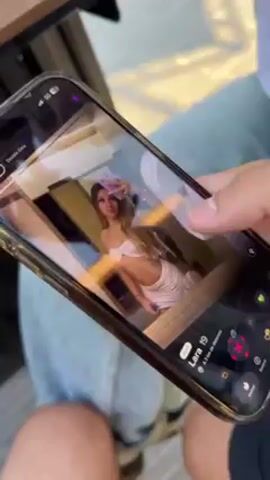 A close-up shot shows a person swiping through profiles on a dating app on their smartphone. The phone is held horizontally, and the person's thumb is seen swiping left and right to dismiss or like different profiles of women. The profiles include photos, ages, and locations of the women. The background shows the person is sitting at a table outside, with another person's leg visible next to them. The overall lighting is bright, suggesting it's daytime.