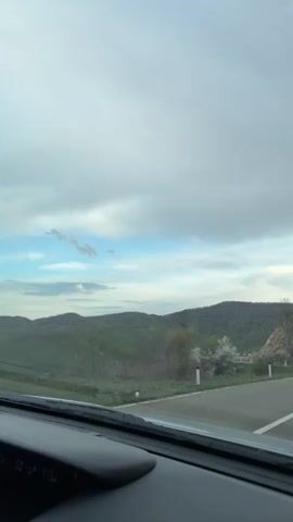 The video shows a car driving on a winding mountain road. The sky is overcast, but there are patches of blue sky. The road has a white dividing line and guardrails. There are trees and hills on either side of the road. Another car is parked on the side of the road. The camera is mounted inside the vehicle, likely on the dashboard.