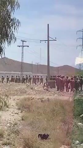 The video captures an outdoor scene in Khuzdar, Pakistan, under a clear, bright, sunny sky. Two distinct groups of uniformed individuals are visible on a dirt road or open ground.

On the left, a group of individuals wearing darker uniforms (possibly green or brown) and helmets is lined up in a formation, appearing somewhat stationary or defensive. Some in this group seem to be holding shields or barricades. A white vehicle is partially visible behind them.

On the right, another group of individuals, clad in reddish-maroon uniforms and helmets, is also in a formation. This group appears to be moving or advancing towards the center, with some members also holding what resemble shields.

Several individuals are positioned in the space between these two main groups, suggesting an interaction, confrontation, or drill is taking place. The background features arid hills or mountains, numerous utility poles with power lines, and a large metal electrical transmission tower on the far right. The immediate foreground shows dry, sparse vegetation and dusty ground. There is no visible text in the scene. The overall setting is rural or semi-urban, characterized by the rugged terrain and visible infrastructure.