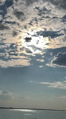 The video shows a view of a body of water with a horizon line in the distance. Above the water is a sky filled with clouds. The sun is partially visible through the clouds, casting bright rays. The clouds are a mix of white and gray, suggesting a dynamic weather pattern. The water appears calm with reflections of the sky and sun on its surface. The overall impression is of a natural landscape scene, possibly taken during daylight hours.