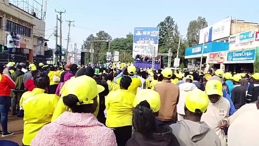 A large crowd of people, many wearing yellow hats, are gathered outdoors in a street. A stage with a large banner displaying text and images is set up at the end of the street, and people are visible on the stage. There are buildings on either side of the street, some with signage. The sky is clear and sunny. The overall impression is of a political rally or public event.