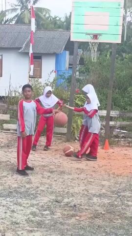 Three children, two girls and one boy, are playing basketball on a dirt court. The boy, wearing a gray and red tracksuit, holds a basketball. The two girls, wearing hijabs and red and gray tracksuits, are standing near him. A basketball hoop is visible in the background. The area appears to be outdoors, with trees and houses in the distance. The sky is overcast. The children are interacting with each other and the basketball.