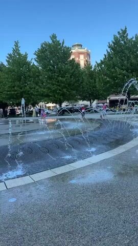 The video shows a public water fountain attraction with jets of water shooting upward. The fountain is circular, with a dark grey surface surrounded by a lighter grey border. Multiple jets of water are active, creating arcs of water. Several people, including children, are visible playing in the fountain. One boy in a blue shirt and black shorts is running through the water jets. Trees surround the area, and a building with a rounded tower is visible in the background. The sky is blue with a few clouds. The overall setting appears to be a park or town square on a sunny day.