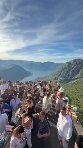 A large crowd of people are gathered on a wooden platform built on a rocky mountainside overlooking a bay with a city nestled along its coast. The sky is bright blue with wispy white clouds. Many people are dressed in summer attire, and some are holding drinks. The scene appears to be a party or social gathering.