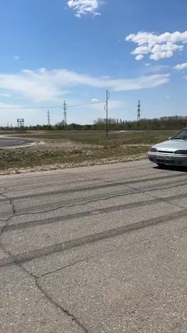 The video shows a light grey Lada sedan, a Russian car, parked with its driver's side door open on a paved road. A man in the driver's seat is visible. The road has multiple skid marks on it. The sedan has aftermarket wheels. The background features a grassy, flat landscape dotted with utility poles and an area fenced off as a racetrack. In the distance, there's a small hill covered in trees. As the car drives away with its door still open, a white SUV and a darker truck are seen in the distance. A small group of people stand by a fence. The day is sunny with a blue sky and scattered clouds. The car bears a Russian license plate. Rap music is playing.