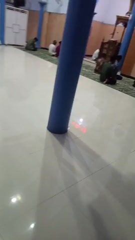The video shows the interior of a mosque. Several men are seated on the floor, facing towards the front of the mosque where a pulpit is visible. Some men are bowing or in prayer. The walls are light brown, and there are blue columns supporting the ceiling. A digital display board showing prayer times is mounted above an archway. The floor is tiled with a light-colored, glossy material. The perspective is from a low angle, looking across the floor and up towards the front of the mosque.