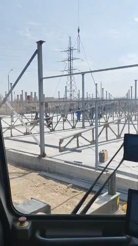 The video shows a construction site from the perspective of inside a crane. In the foreground are the interior workings of the crane's cabin, including a monitor and control panels. The main view through the windshield reveals a large, open construction site with a concrete foundation. Metal beams and support columns are being assembled, creating a framework. Several construction workers, dressed in blue work clothes, are present on the site, some of whom appear to be welding or fastening the metal structures together. A large crane is visible in the background, lifting a piece of equipment or material into position. An electrical transmission tower stands prominently in the distance, with power lines stretching across the sky. The ground outside the foundation is dusty and barren. The sky is clear and sunny, creating bright lighting across the scene.