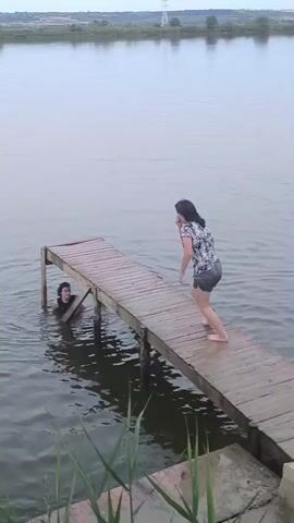 A young woman in a black and white patterned shirt and dark shorts jumps off a wooden dock into a body of water. She lands with a splash, creating a large ripple. A colorful inflatable ring floats nearby in the water. The woman surfaces and appears to be laughing or calling out. The environment is a lake or calm river with reeds in the foreground and a tree line in the background under an overcast sky.