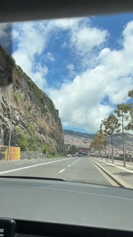 The video shows a view from inside a car driving on a road next to a cliff and a pedestrian walkway. A few trees line the walkway. Across the road, a tall, rocky cliff face rises, with some greenery growing on it. Further down the road, there's a red brick building. In the background, a densely populated area is visible on the side of a hill. The sky is blue with scattered white clouds. The weather appears to be sunny. There are a few other vehicles on the road and some people walking on the walkway.