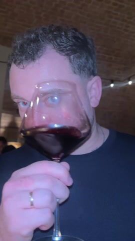 A man with short curly brown hair and a light beard is shown up close, taking a drink of red wine from a large, clear wine glass. The man is wearing a black shirt and a gold ring on his left ring finger. The background is out of focus but appears to be inside a building with a brick ceiling. String lights are visible in the background. The camera focuses very closely on the man's mouth as he drinks the wine.