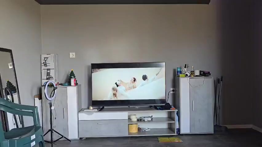 Here is a description of the video.

The video shows a living room. In the center is a TV displaying a music video featuring a woman in an elaborate outfit and two people in white clothing kneeling. To the left of the TV is a white and gray cabinet with a Christmas tree and a ring light on top. To the right is a similar cabinet cluttered with various items. The wall behind the furniture is gray.