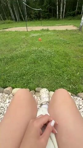 The video begins with a close-up shot of a person's legs and feet, partially obscured by a white towel. The person is sitting on a surface covered with white pebbles. In the background, a green lawn is visible, with a volleyball net and an orange ball in the distance. Two white slippers are placed on the pebbles near the person's feet. The camera then pans up to reveal the person's bare feet, which are also on the pebbles. The scene then transitions to a wider shot showing the entire yard, with trees in the background. The person is still seated, with the white towel still in their lap. The orange ball is visible on the lawn, near the volleyball net. The video ends with another shot of the person's legs and feet.