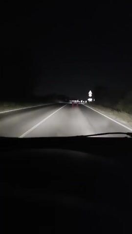 The video shows a first-person view from inside a car driving on a road at night. The headlights of the car illuminate the asphalt road, which appears wet. White lane markings are visible on the left side of the road. In the distance, the taillights of another car are visible, along with some streetlights. Several road signs are also visible on the right side of the road, including a triangular sign with a red border and a white background, a circular sign with a red border and a white background, and a square sign with a white background and black symbols. The environment is dark, with the exception of the car's headlights and the illuminated signs and taillights.