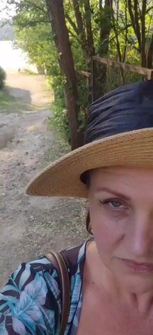 The video shows a close-up of a woman wearing a straw hat, looking towards the camera. The background is out of focus, but it appears to be a natural setting with trees and a body of water in the distance. A dirt path leads downhill towards the water. There are some colorful playground objects visible near the path. The woman's expression is serious. The lighting suggests it is daytime, possibly late afternoon.