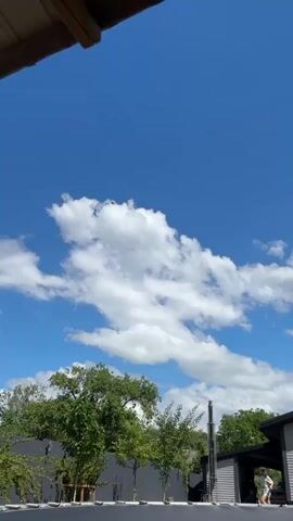 A drone is flying in the sky, it is a bright sunny day with a clear blue sky and some fluffy white clouds. The drone is a small dark object against the sky. In the foreground, there is a wooden overhang, and the tops of green trees can be seen below.