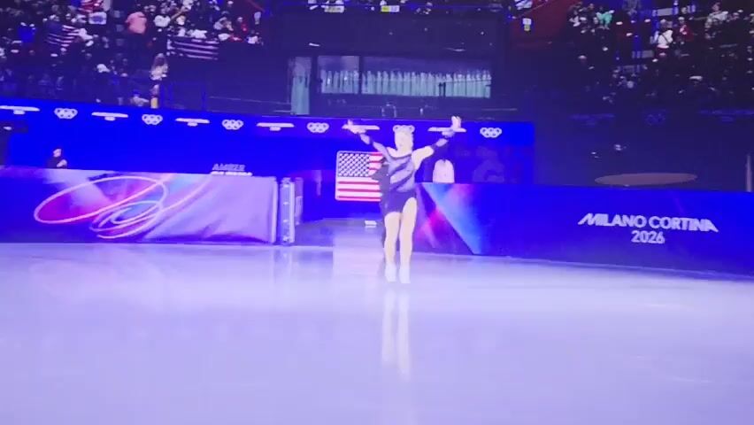An ice skater stands in the center of an ice rink, her arms raised in a triumphant pose. She is wearing a dark blue skating dress with sheer panels and white skates. Her blonde hair is pulled back. Behind her are event banners for "MILANO CORTINA 2026" and an American flag. A crowd is visible in the background. The lighting is bright and focused on the center of the rink.
