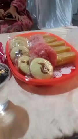The video shows a close-up view of a tray of Indonesian snacks placed on a table covered with a light-colored tablecloth. The snacks include round, pale yellow cakes, a pile of pink, granular confectionery, and several rectangular, golden-yellow pieces. The snacks are partially covered in clear plastic wrap. To the left of the tray is a clear plastic cup filled with a clear liquid and topped with a black lid. Beside the cup is a package of pink and white tissues. In the background, a person wearing a pink and red patterned outfit is visible, holding a mobile phone. There are also white draped cloths in the background, suggesting an event setting. A jar filled with snacks is partially visible on the far left. The lighting is dim, with shadows cast on the tablecloth.