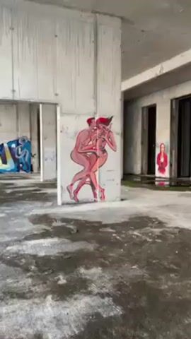 The video shows the interior of a derelict concrete structure. Graffiti and paintings are on the walls. One painting shows two figures entwined. A person wearing dark clothes and a hood walks to the right side of the frame and disappears from view. The floor is concrete and has areas of discoloration. There are open doorways, and structural pillars are visible.