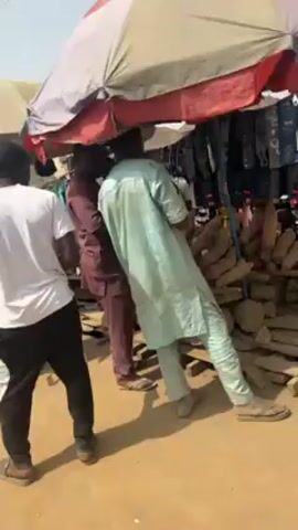 The video shows an outdoor marketplace on a sunny day. Several people are present, some carrying goods on their heads. There are makeshift stalls with umbrellas shading the vendors. Some stalls display clothing for sale. Other stalls have piles of vegetables like tomatoes and leafy greens. The ground appears to be a mix of dirt and paving stones. In the background, there are houses along the street.