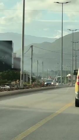 The video shows a road with traffic. A black Volkswagen Touareg is visible from the rear. A yellow lane divider is painted on the road. There are streetlights lining the road, with mountains visible in the background under a cloudy sky. A bus is driving in the distance. A restaurant sign that reads "GRILL RESTORAN" is visible on a building to the side of the road. Construction work is underway on the side of the road.