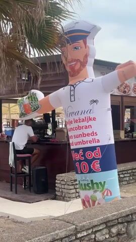This media file captures an outdoor scene, likely at a beachside establishment in Bečići, Montenegro. The primary focus is a tall, inflatable tube man dressed as a sailor, complete with a sailor's hat, blue eyes, and a red beard. He is holding a beer in one hand and his other arm is raised in a wave. His shirt displays advertising text and imagery.

Behind the inflatable figure, a counter or bar area is visible with a few people. One person, wearing a white t-shirt and black shorts, is seated at the counter and appears to be interacting with someone behind it. Another person, also in a white t-shirt, is working behind the counter, holding a cocktail shaker. A Coca-Cola sign is visible on the counter.

The background suggests a casual, possibly tourist-oriented setting, with wooden structures and glimpses of what might be beach umbrellas and palm trees. The lighting indicates it is daytime, with overcast or bright sky.

The inflatable man's shirt features text that appears to be in a Slavic language. The visible text includes "Nirvana", a palm tree graphic, and phrases that seem to relate to "sunbeds" and "umbrella" rental. A prominent price is displayed as "Već od 10 €" (Already from 10 €). The bottom of the inflatable depicts ice cream cones and a beach umbrella, suggesting an offering of refreshments.

The overall mood of the scene is relaxed and commercial, typical of a summer vacation spot. The inflatable figure is a whimsical advertisement, designed to attract attention and convey a sense of fun and affordability for the services offered.
