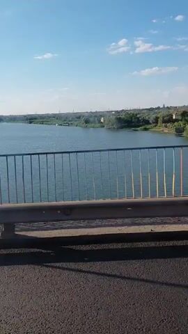 The video shows a view from a bridge overlooking a body of water, likely a river or lake, with a shoreline dotted with trees and houses. The sky is clear blue with scattered white clouds, suggesting a pleasant day. The time of day appears to be late morning or early afternoon due to the bright sunlight and the shadows cast on the road in the foreground.

The bridge railing, painted with vertical yellow and blue stripes, is prominent in the foreground. Beyond the railing, the water stretches out, reflecting the sky. The opposite bank is covered in lush green trees, with several houses visible among them, some featuring red-tiled roofs. The houses seem to be part of a small village or settlement nestled by the water.

In the distance, the landscape appears somewhat flat, with more vegetation and some structures that could be industrial or residential buildings. The overall scene evokes a sense of peace and tranquility, characteristic of a rural or semi-rural setting.

The location is identified as Maiaky, Ukraine. The weather is sunny and clear, contributing to the serene atmosphere. No people are visible in the video, nor is there any visible text. The activity taking place is simply the act of traveling across the bridge, offering a scenic view.