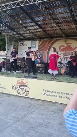 A musical band performs on an outdoor stage. There are six people on stage. Two men play guitars, one man plays an accordion, and another man plays drums. Two women sing, one in a red dress and one in a black dress. The stage has a metal frame and a corrugated roof. A yellow banner with the text "Крутий Заміс" and a clay pot logo is displayed in front of the stage. Another banner with Ukrainian text and a QR code is visible in the background. A person's arm wearing a striped shirt is visible in the bottom right corner, suggesting an audience member. The ground in front of the stage is made of light-colored pavement or concrete.