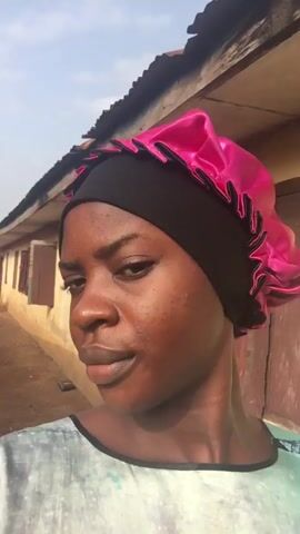 A young woman with dark skin is captured in a close-up, selfie-style video outdoors in Isieke, Nigeria. She wears a black underscarf topped with a vibrant pink, ruffled satin bonnet and a light-colored, patterned top.

She poses for the camera, shifting her gaze between directly at the lens and looking slightly off-camera. Throughout the video, she casually touches her bonnet or head with her right hand. For a brief moment, she holds a small bunch of keys near her head. Her facial expression remains mostly neutral and composed, occasionally appearing thoughtful or serene.

The background changes slightly, initially showing a weathered, yellow or light brown building with closed wooden shutters under a clear sky, suggesting daytime. Later, a rusty metal gate and other distant buildings, along with clothes hanging to dry, become visible, indicating a residential or community area. There is no visible text.