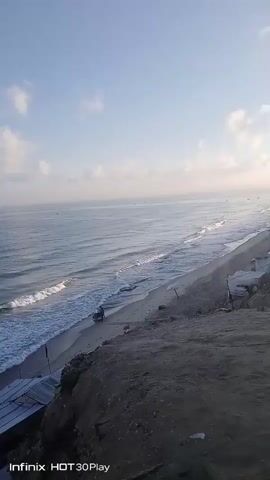 A high-angle, wide shot captures a view of a serene ocean with gentle waves lapping against a sandy beach. The sky is a soft blue with scattered white clouds. Along the edge of the beach, there are several makeshift shelters with striped canvas roofs. A person is seen riding what appears to be a motorcycle or scooter on the sand near the water's edge. The foreground shows a rocky, elevated cliff face. The bottom corner of the video displays a watermark indicating it was recorded on an "Infinix HOT 30 Play."
