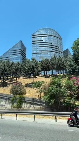 A person on a red scooter wearing a white helmet and a black shirt rides along a road. In the background, two modern glass buildings stand on a hill covered in trees and dry grass. A white van partially obstructs the view in the foreground. The sky is clear and blue.