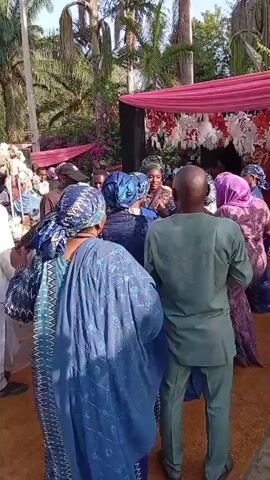 The video shows a group of people dancing at what appears to be an outdoor event. Most of the women are wearing similar blue patterned dresses and headwraps, while a man in a green suit stands at the edge of the group. They are gathered near a stage-like structure decorated with pink and white fabric. Palm trees are visible in the background. The surface underfoot appears to be sand. People are talking and laughing. One woman is filming with her phone.