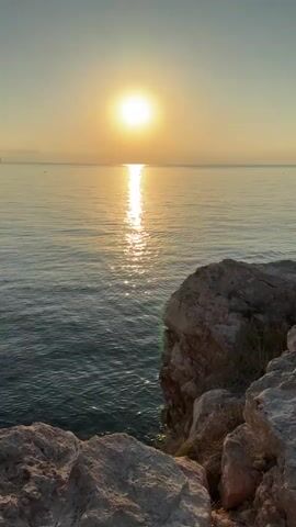 The video shows a sunrise over the ocean, with the sun appearing as a bright, glowing orb just above the horizon. The sun's reflection creates a shimmering path across the water's surface. The foreground of the scene is dominated by large, textured rocks, some of which are covered with dry grass. The water appears calm, with gentle ripples. In the far distance, a faint outline of land or buildings can be seen. The overall mood is peaceful and serene.