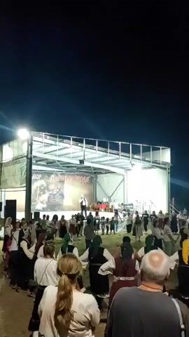 A nighttime video shows a traditional festival in an open field, with a stage set up for live music. Performers are on stage, and a crowd is gathered below, with many people participating in a traditional dance, forming a large circle and holding hands. The background is dark, indicating night, with bright lights illuminating the stage and the immediate area.