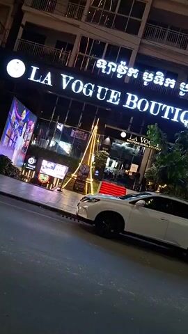 The video shows a white Lexus SUV parked on a street at night. The SUV is in the foreground and is angled slightly towards the right of the frame. Behind the SUV is a building with a large illuminated sign that reads "LA VOGUE BOUTIQUE HOTEL & CASINO" in English and Khmer. The building appears to be a hotel and casino. There are trees and greenery in front of the building, and some motorcycles are parked on the sidewalk. The street is paved and appears to be in a city. The lighting suggests it is nighttime or dusk.