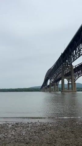 The video shows a steel bridge stretching across a body of water. The bridge has a complex structure of crisscrossing metal beams and concrete support pillars. The water is gray, with small waves rippling across its surface. In the background, a line of green trees and rolling hills is visible under a cloudy sky. The foreground features a muddy shoreline.