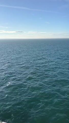 This is a wide shot of a vast, calm ocean under a bright, clear blue sky. The horizon line is sharp and distinct, separating the deep blue of the sea from the lighter blue of the sky. A few wispy white clouds are scattered across the sky, hinting at a pleasant day. The water is a rich teal color, with gentle ripples creating a textured surface that reflects the sunlight. In the distance, the sea appears almost flat, merging seamlessly with the sky.

The scene is serene and expansive, evoking a sense of peace and tranquility. The lighting suggests it is daytime, likely late morning or early afternoon, given the brightness of the sun. There are no people or man-made objects visible, emphasizing the natural beauty and solitude of the ocean. The water's surface is mostly undisturbed, with only subtle waves suggesting a light breeze. It's a classic seascape, presenting the immense power and beauty of nature.