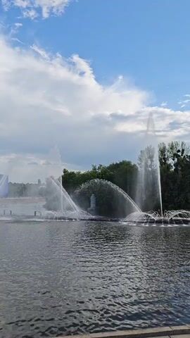 The video shows a wide fountain spraying water into the air over a body of water. Trees are visible in the background, and the sky is blue with scattered clouds. The water from the fountain creates a misty effect.