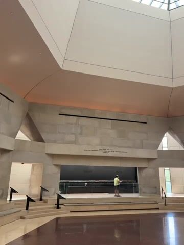 A wide shot pans across the interior of a large, modern building with a domed ceiling and skylights. The walls are constructed of large, light-colored stone blocks. In the center of the room, a person wearing a yellow shirt and dark shorts stands before a wall with numerous small lights arranged in rows. They appear to be looking at the lights. To the right of this display, there is a large, dark rectangular plaque with text engraved on it, and a flame burning in front of it. The floor is a reflective, dark red material. The overall impression is of a solemn memorial or museum.