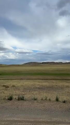 This media file captures a vast, open landscape with a cloudy sky. The foreground features a gravelly dirt road and sparse, dry vegetation. Beyond the road, a wide expanse of grassland stretches towards a line of low, undulating hills in the distance. The grassland is a mix of dry, yellowish-brown grass and patches of green, suggesting varied terrain or recent rainfall in different areas.

The sky is dynamic, filled with a mix of dramatic clouds. Large, white, fluffy clouds are interspersed with darker, more ominous storm clouds. Shafts of light are visible breaking through the clouds, creating streaks of illumination on the distant landscape, hinting at the possibility of rain. The overall impression is one of a wide-open, natural environment under a changeable sky. There are no visible people or man-made objects other than the dirt road. The scene evokes a sense of solitude and the raw beauty of nature. The lighting suggests it is likely daytime, possibly late afternoon given the dramatic cloud formations.